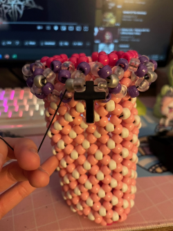 Photographed in the middle is an X-cuff that is light pink and white, on the top there is a crown that consists of purple, silver, and frosty clear beads, there is a black cross on it as well. Behind this is a desk with a keyboard, mouse, and computer screen with YouTube displayed on it.