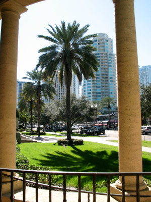 This is the outside of the St. Pete of Museum of Fine Arts, there are two columns and a black fence, what goes past that are full palm trees, sidewalks, and buildings that reach high into the sky.