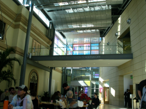 This is a view of the lobby in the St. Pete Museum of Fine Arts, there is more colorful windows that reflect light onto the walls. There is a bridge in the middle of the image, underneath that bridge are guests sitting and chatting with each other.