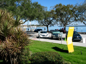 Outside, in the back of the St. Pete Museum of Fine Arts is a parking lot, there are cars parked in it, behind it is the beautiful blue ocean, there is a yellow sign that points to the museum, framing this image are shrubbery and trees.