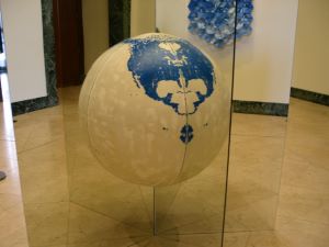 In a white corridor, there is a globe in the middle of the room, there are two mirrors that intersect it in an X shape. The globe is white, grey, and blue. There is an art piece on the wall as well, it is multiple ceramic cups that are blue and white.