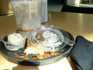 On this table is a plate that has a finished coffee drink, cold, un-spreadable butter in a little plastic cup, to its right the lid is on top a mostly-eaten croissant, and there is a fork next to it as well. Next to said plate is a black face mask. If you are reading this, eat before you go to a museum.