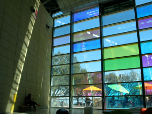 There are two walls that meet, on the right is a wall made entirely of windows, some of them are green, yellow, grey, blue, and pink. The left wall is a paneled beige and there is someone sitting against that wall. In front of the window wall are beanbag chairs. 